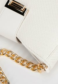 Bílá požární taška s texturovaným povrchem, zlatým řetízkem a zlatými doplňky. Zavírání má detail obdélníkového designu.