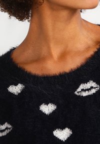 Black fuzzy sweater featuring white knitted heart and lip patterns, with a wide neckline and soft texture. Close-up of neckline area.