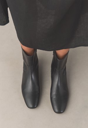 Person trägt schwarze Lederstiefel bis zur Mitte der Wade und einen schwarzen Rock, steht auf einem hellgrauen Boden.