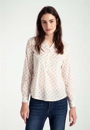 Blouse blanche avec des pois marron clair, manches longues, patte de boutonnage et coupe décontractée. Portée avec un jean en denim foncé.