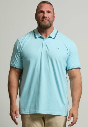 Erwachsener Mann mit Bart, der ein hellblaues Polohemd und beige Hose trägt, steht vor einem einfarbigen grauen Hintergrund und schaut nach vorne.