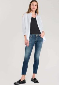 Camiseta de tirantes negra debajo de una camisa blanca de botones abierta, combinada con jeans oscuros ajustados y mocasines negros, de pie sobre un fondo liso.