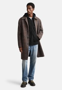 Brown plaid overcoat with large front buttons, worn over a black zip-up hoodie. Light blue jeans and dark shoes with green accents complete the outfit.