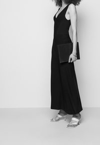 Svart ärmlös jumpsuit med V-ringning, som har en figurnära överdel och vida ben. Modellen håller en svart mapp och bär vita sandaler med remmar.