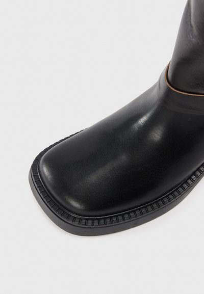 Botte en cuir noir avec un bout arrondi, présentant une texture lisse et une semelle en caoutchouc à rainures. Le design comprend un accent discrètement cousu.
