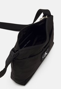 Musta olkalaukku, joka on valmistettu tekstuurillisesta kankaasta. Siinä on pääasiallinen vetoketjullinen lokero, säädettävä olkahihna ja etupuolella oleva "ck"-logolla varustettu applikaatio.