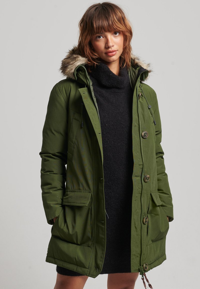 Grüne gepolsterte Parka mit einer Kapuze aus Kunstpelz, Vorderknöpfen und seitlichen Taschen. Getragen über einem dunklen Pullover, der ein lässiges Design zeigt.