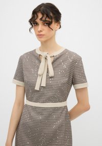 Šaty s krátkým rukávem v béžovém a hnědém vzoru houndstooth, s detailem zavazovacího límce a jemnými třpytivými akcenty na látce.
