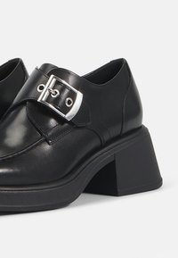 Pantofi din piele neagră cu toc gros, având o curea lată și detalii cu cataramă argintie lucioasă. Textură netedă cu cusături curate.