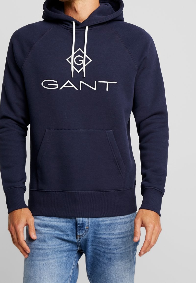 Felpa blu navy realizzata in misto cotone, con grande logo GANT bianco, cappuccio con cordino e tasca frontale a marsupio.
