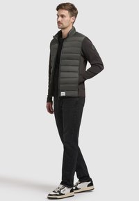 Mørkegrøn quiltet vest med høj krave, båret over en sort sweater, parret med sorte jeans og sneakers med hvide detaljer.