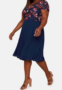 Námornícky A-liniový šaty s květinovou krajkovou živůtkou a krátkými rukávy, s výstřihem do V a splývavou sukní. Doplnené nahými sandálkami na hrubém podpatku.