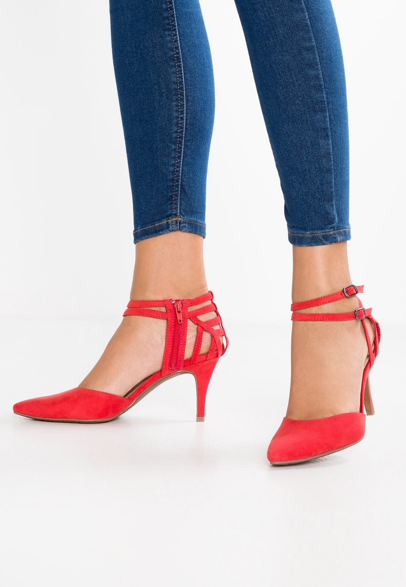 Pieds chaussés de talons hauts rouges avec des brides à la cheville, assortis à un jean skinny bleu sur fond blanc.