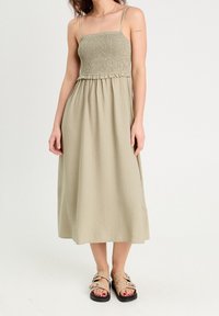 Vestido verde oliva claro con cuerpo fruncido, tirantes finos y una falda en línea A que fluye. Textura notable y detalles de diseño de longitud media.