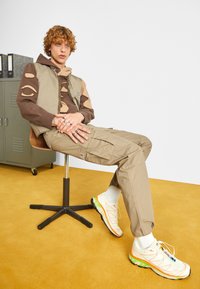 Beiga cargobyxor, avklippt brun hoodie med utskurna detaljer, och vita sneakers med orange och gröna accenter. Sitter på en brun snurrstol.