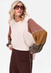 Sweter o ribbowanym kremowym korpusie z wielokolorowymi balonowymi rękawami w kolorach burgundowym, musztardowym i granatowym. Połączony z brązowymi spodniami w paski.