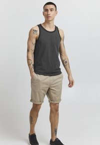 Débardeur gris foncé avec un col noir, associé à un short beige retroussé et des baskets noires. Tatouages notables visibles sur les bras.