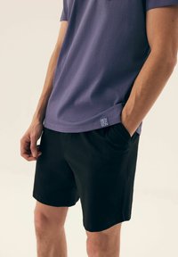 Fioletowa koszula z krótkim rękawem o prostym wykończeniu, w komplecie z czarnymi szortami z kieszeniami. Mała metka marki widoczna w pobliżu krawędzi.