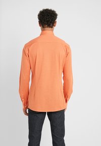 Camisa de manga larga en naranja brillante, confeccionada en tejido suave. Presenta un cuello puntiagudo y puños abotonados. Combinada con pantalones de mezclilla oscuros.