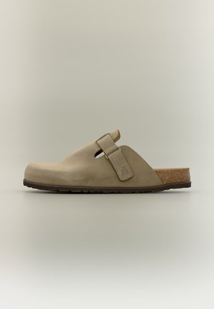 Sabot beige en suède à enfiler avec sangle réglable, semelle intérieure en liège et semelle extérieure en caoutchouc noir, présenté de profil sur fond neutre.