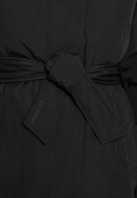 Casaco com cinto preto apresentando uma textura suave, com um cinto largo e detalhes costurados que exibem o logótipo da Calvin Klein.