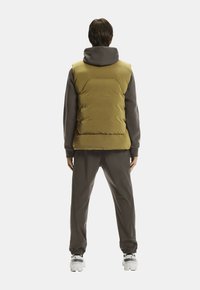 Person steht mit dem Rücken zur Kamera und trägt eine senffarbene Steppweste über einem dunkelgrauen Hoodie, passende Jogginghosen und weiße Sneakers vor einem weißen Hintergrund.