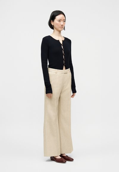 Kvinne iført svart knappet cardigan, beige vide bukser og brune flate sko, stående mot en ensfarget lys bakgrunn.