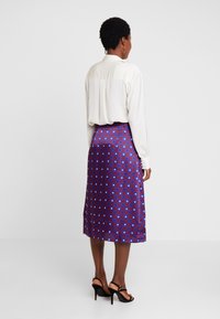 Jupe midi en satin bleu et rouge avec des motifs géométriques ; texture lisse, taille haute et fente sur le côté. Associée à un blouse blanche.