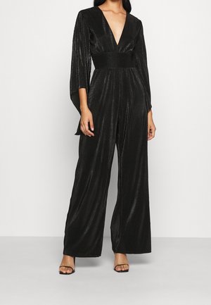 Femme portant une combinaison noire en tissu texturé avec jambes larges, un décolleté en V profond et des manches trois-quarts cape, associée à des escarpins noirs à bout ouvert.