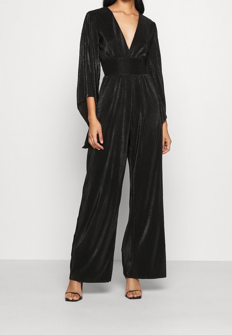 Femme portant une combinaison noire en tissu texturé avec jambes larges, un décolleté en V profond et des manches trois-quarts cape, associée à des escarpins noirs à bout ouvert.