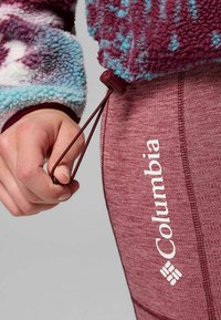 Sudadera de forro polar con patrón multicolor y cordón ajustable, combinada con leggins burdeos que presentan un logo blanco y una textura elástica.