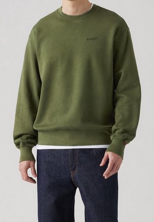 Mand iført en olivengrøn crewneck sweatshirt med lille logo på brystet, kombineret med mørkeblå jeans og en hvid undershirt.