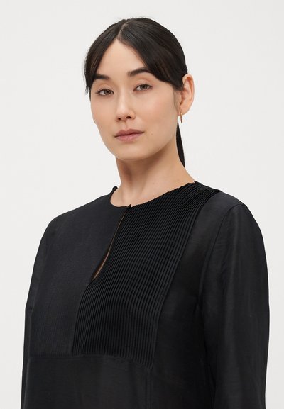 Bluza neagră cu detalii cu pliuri pe panoul din față, decolteu rotund și material ușor. Are o deschidere mică tip cheiță.