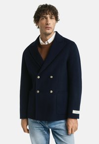 Navyblauwe dubbelbreasted wollen jas met zilveren knopen, met een stuikrevers en twee voorzakken, gedragen over een bruine trui en een witte shirt.