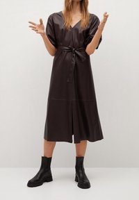 Vestido de faux cuero marrón con escote en V, mangas cortas, cinturilla ceñida y abertura en el centro delantero. Combinado con botas negras hasta el tobillo.