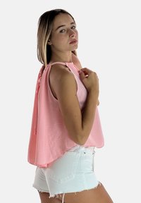 Top sin mangas rosa con escote halter y detalle de parte posterior fruncido, combinado con pantalones cortos de mezclilla blanca desgastados. Textura de tela suave.