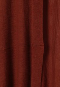 Tessuto a maglia color ruggine con una texture liscia, caratterizzato da una trama allentata e leggere variazioni del motivo. È visibile un particolare della cucitura.