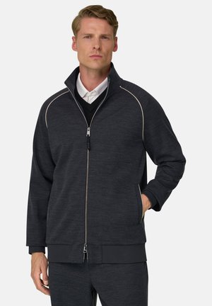 Mand iført en mørkegrå lynlåstrøje over en sort sweater og hvid skjorte med krave, stående med den ene hånd i lomme på jakken.