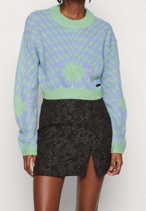 Kvinde iført en kort grøn og lavendel mønstret sweater med en stjerneform og et sort blomstertekstureret skørt med en frontslids.