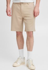 Shorts beiges en tissu lisse, avec une fermeture à bouton et des poches avant. Associés à des chaussettes blanches et des baskets crème.
