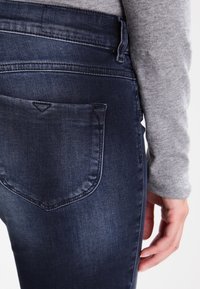 Tmavě modré džíny z denimu s viditelnou zadní kapesou. Látka má hladkou texturu s jemným vyblednutím. Jsou patrné detaily zipu a šití.