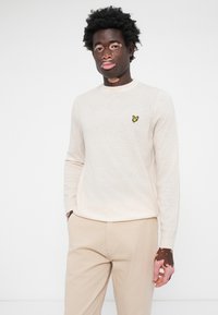 Pull en tricot beige clair à manches longues et col rond, avec un petit emblème de papillon jaune sur la poitrine. Porté avec un pantalon beige.