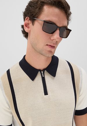 Giovane uomo che indossa occhiali da sole scuri e una polo lavorata a maglia beige con strisce verticali nere e marroni.