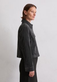 Czarna kurtka z corduroy z kołnierzykowym dekoltem, pojedynczą kieszenią na piersi i guzikami na mankietach. Ma krótki krój i teksturowany materiał.