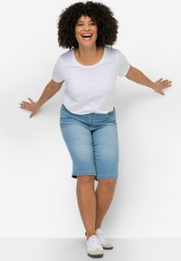 T-shirt bianco a maniche corte abbinato a pantaloni shorts in denim azzurro chiaro fino al ginocchio. Il modello indossa sneaker bianche con dettagli grigi.