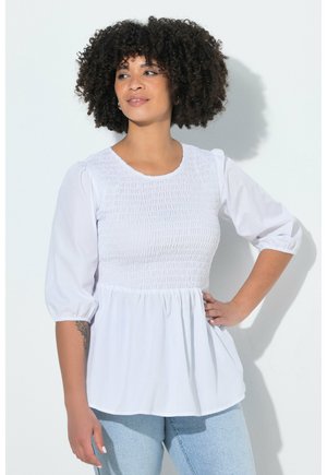 Kvinde med krøllet hår iført en hvid smock-bluse med albuelange ærmer og lyseblå jeans, stående med den ene hånd på hoften.