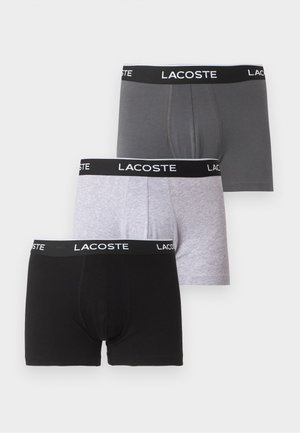 Homme à la peau foncée debout pieds nus sur un fond blanc, portant un boxer gris Lacoste, un bras croisé sur la poitrine.