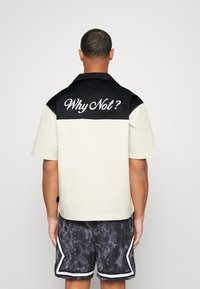 Camicia a maniche corte con un pannello superiore nero e un fondo avorio. Presenta un ricamo sulla schiena con il testo "Perché no?".