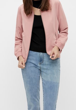 Femme portant une veste bomber rose clair zippée, un haut côtelé noir et un jean bleu clair, les mains près de la fermeture éclair et des poches de la veste.