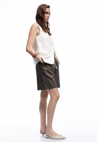Weißes ärmelloses Oberteil mit Knöpfen, kombiniert mit olivgrünen Shorts; Model trägt helle Slingback-Flats und ein Holzarmband.